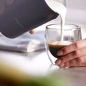 Een hand met een donkergrijze Philips melkopschuimer schenkt melkschuim in een glas espresso met dubbele wand.