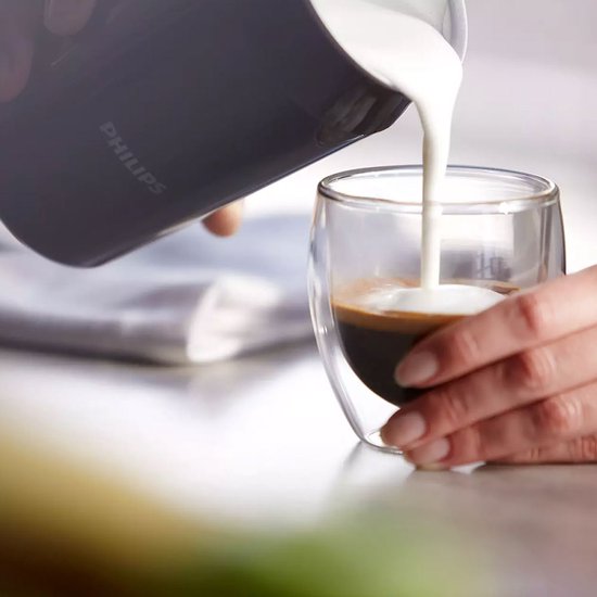 Een hand met een donkergrijze Philips melkopschuimer schenkt melkschuim in een glas espresso met dubbele wand.