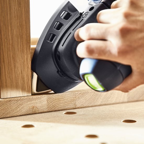 Een hand houdt een zwarte schuurmachine met een groen accent en een puntige schuurzool, die een hoek van hout schuurt.