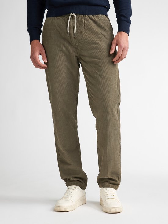 Model draagt een olijfgroene corduroy broek met trekkoord, marineblauwe trui en witte sneakers.