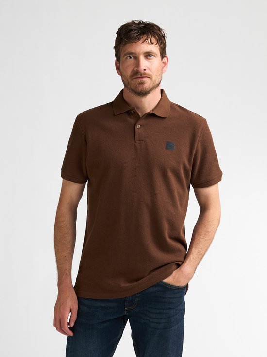 Man in een bruin katoenen poloshirt met korte mouwen en spijkerbroek, kijkend naar links.