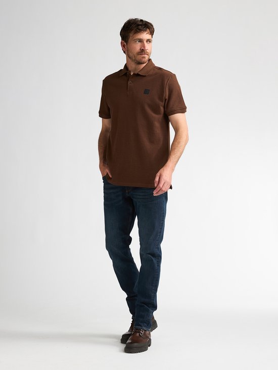 Man in een bruin katoenen poloshirt met korte mouwen en spijkerbroek, staand.