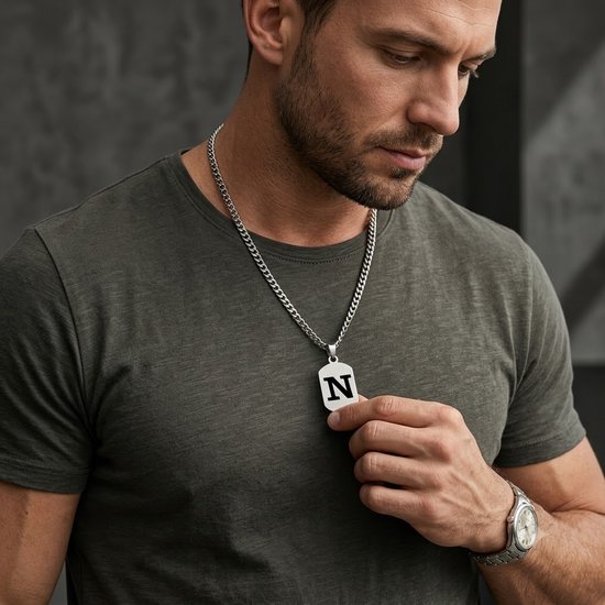 Man met donkergroen T-shirt draagt een zilverkleurige schakelketting met rechthoekige hanger met de letter N.