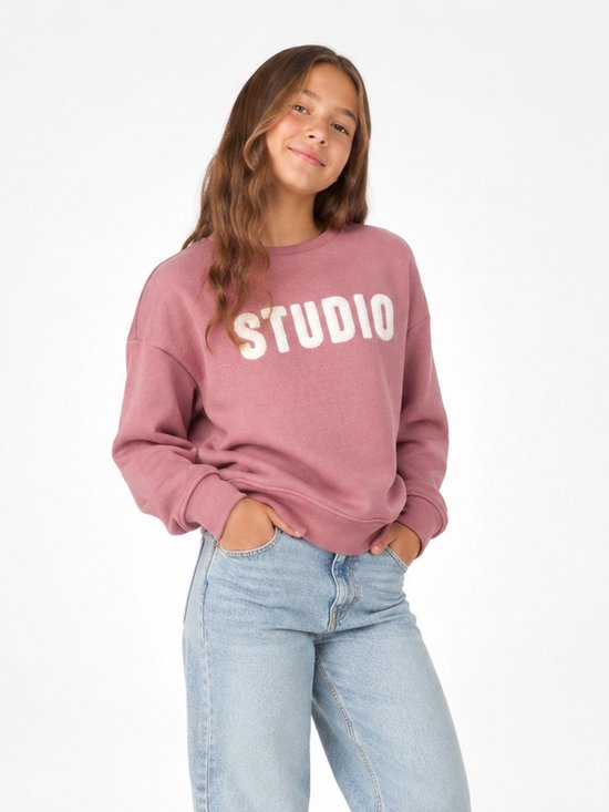 Meisje met lang bruin haar in een roze 'STUDIO' sweater en spijkerbroek.