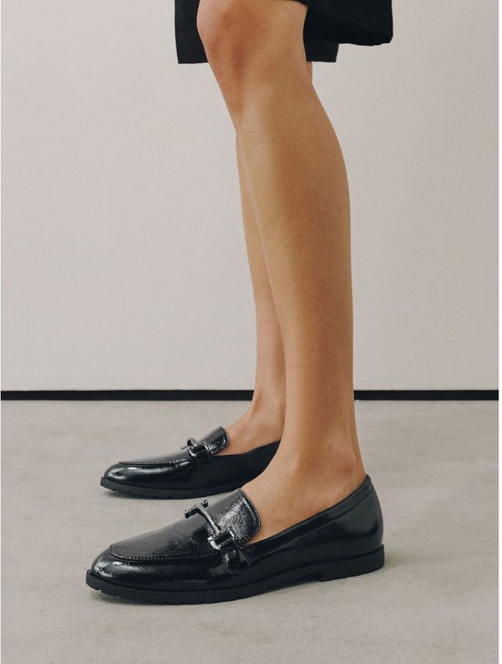 Voeten van een vrouw met zwarte glanzende loafers met zilverkleurige gespen.