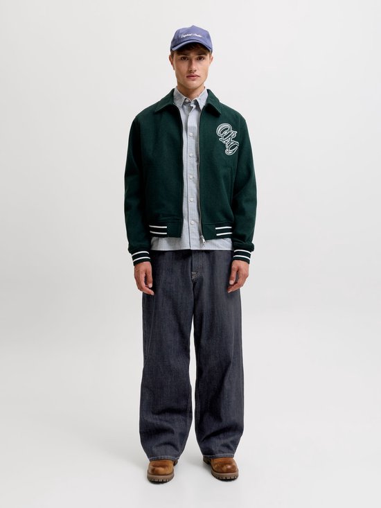 Model draagt een donkergroen jack met logo, geruit overhemd, wijde donkere jeans en bruine laarzen.