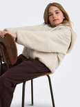 Meisje zit op een stoel en draagt een beige teddy jas, bruine joggingbroek.