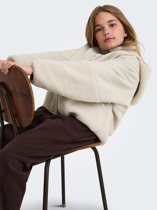 Meisje zit op een stoel en draagt een beige teddy jas, bruine joggingbroek.