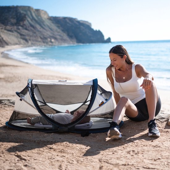 Vrouw op het strand kijkt naar baby in Deryan Baby Luxe campingbedje.