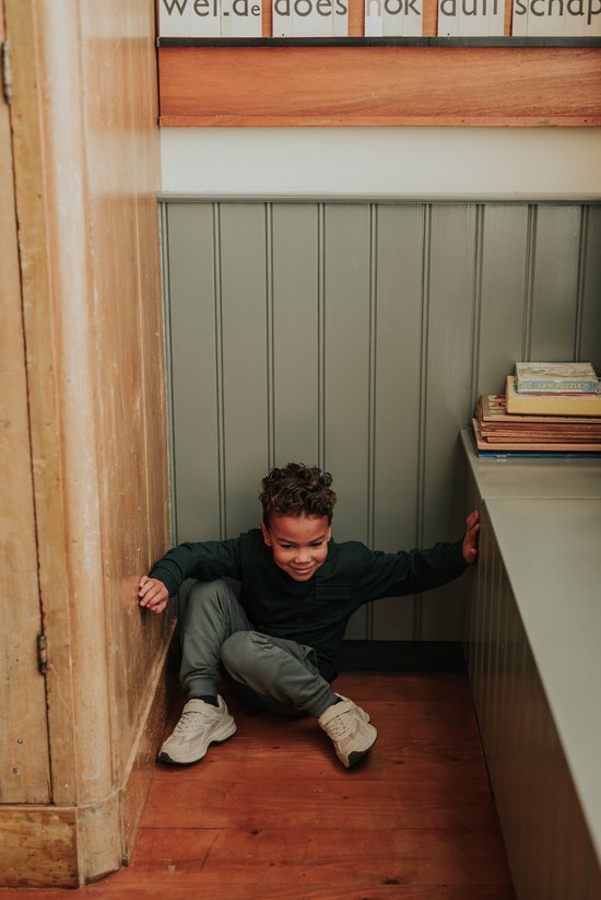 Jongen met krullend haar, gekleed in een donkergroene trui en grijze broek, zit op de houten vloer.