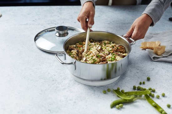 Une personne remue un risotto aux champignons et aux pois dans une casserole argentée avec une cuillère en bois.