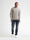 Man in een grijze trui en donkerblauwe jeans, staand op een witte achtergrond.