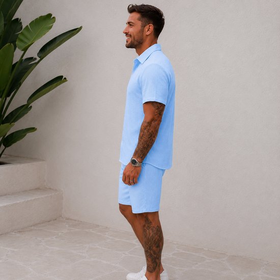 Man, zijaanzicht, draagt lichtblauwe korte mouw blouse en korte broek. Blouse met structuur. Witte sneakers.