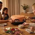Een familie maakt pizza's rond de terracotta Pizzarette pizzaoven met ingrediënten.