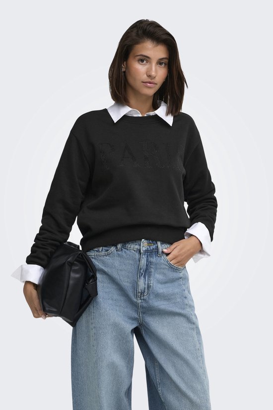 Model in zwarte sweater met "PARIS" opdruk van parels, wit overhemd en jeans.