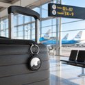 Zwarte AirTag-sleutelhanger aan een koffer bij een luchthaven met vliegtuigen op de achtergrond.