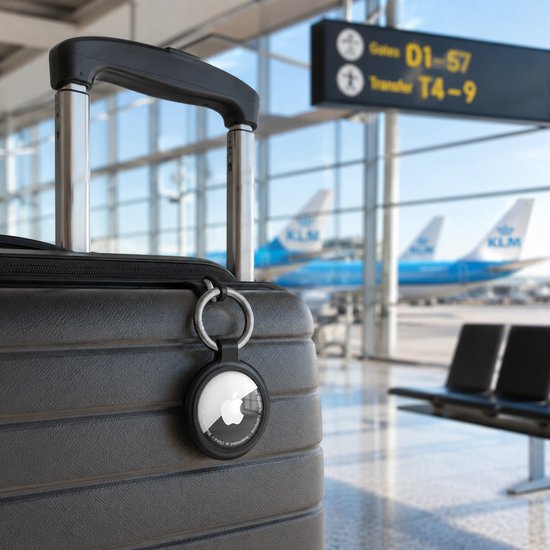 Zwarte AirTag-sleutelhanger aan een koffer bij een luchthaven met vliegtuigen op de achtergrond.