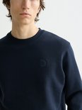 Close-up van een donkerblauwe sweater met ronde hals en een vierkant logo op de borst.