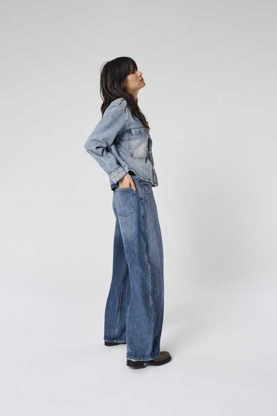 Vrouw in spijkerjack en wijde, middendonkere spijkerbroek met hoge taille. Ze staat met handen in de zakken.