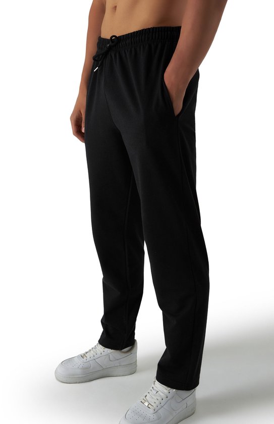 Model in zwarte joggingbroek met trekkoord, witte sneakers en handen in de zakken.