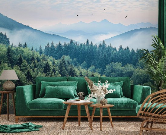 Décoration murale avec montagnes, forêt et brume, au-dessus d'un canapé vert et de tables en bois.