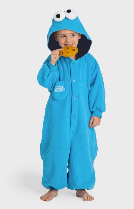 Un garçon porte une combinaison bleue Cookie Monster avec une capuche et des yeux, mangeant un biscuit. Le texte 'COOKIE MONSTER' est sur la poche de poitrine.