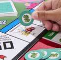 Hand houdt een groene T-Rex token vast boven een speelbord van Monopoly Junior.