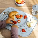 Assiette bleue pour enfant avec imprimé d'animaux et de champignons. Enfant coupant une fraise avec un petit couteau. Doigts d'enfant visibles.