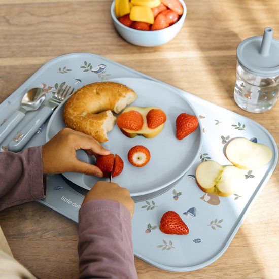 Assiette bleue pour enfant avec imprimé d'animaux et de champignons. Enfant coupant une fraise avec un petit couteau. Doigts d'enfant visibles.