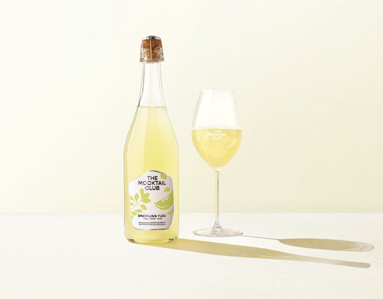 Bubbly alcoholvrije Sparkling Yuzu mocktail van The Mocktail Club in fles en glas. Goudgeel met een groene label.