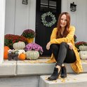 Vrouw in een gele jas zittend op de treden van een huis met pompoenen en bloemen.