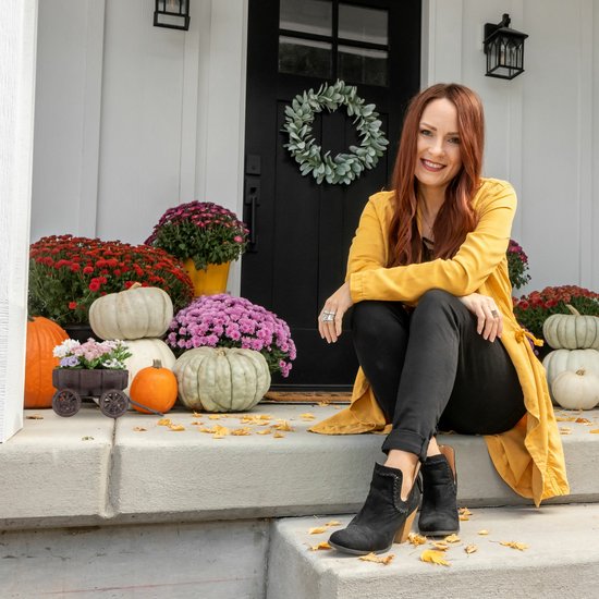 Vrouw in een gele jas zittend op de treden van een huis met pompoenen en bloemen.