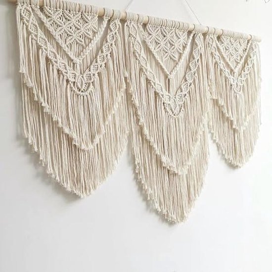 Crèmekleurig macramé wandkleed met drie hangende V-vormige patronen, op een houten stok.