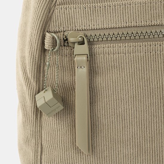 Close-up van een beige ribfluwelen rugzak met een rits, ritstrekker en een vierkant bedeltje met 'Hedgren'.