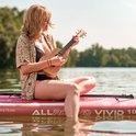Een vrouw met blond haar speelt op een naturel houten concertukelele terwijl ze op een paddleboard zit.