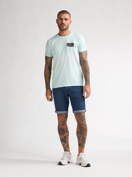 Man met korte mouwen lichtgroen T-shirt met opdruk op de borst en spijkerbroek.