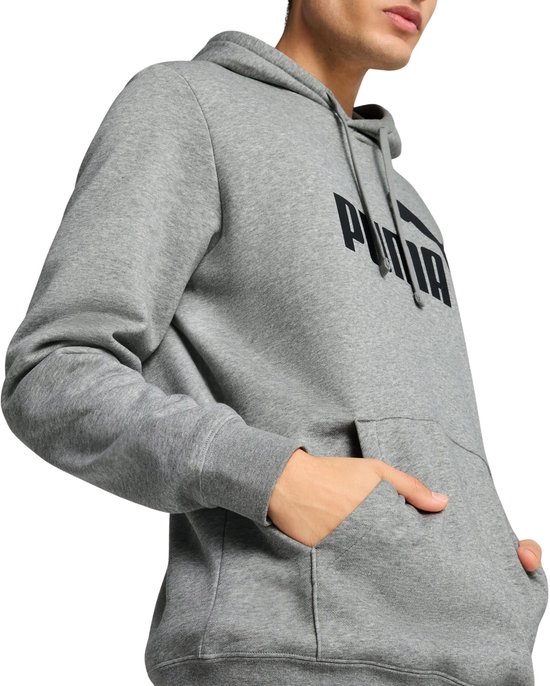 Man in een grijze PUMA hoodie met zwarte opdruk, met de hand in de kangoeroezak.