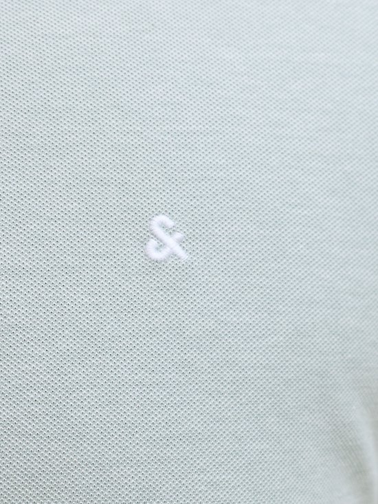 Close-up van het geborduurde '&' logo op een lichtgroen poloshirt.
