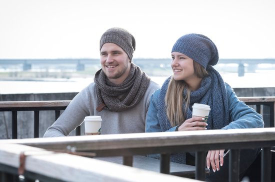 Een stel zit buiten bij een tafel, de vrouw met een blauwe gebreide muts en sjaal, de man met een grijze muts en sjaal.