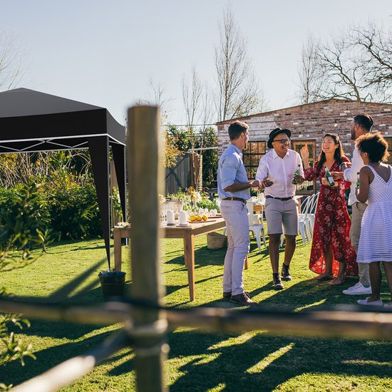 Zwarte partytent in een tuin tijdens een zonnige dag. Mensen staan rond een tafel met drankjes en eten, op een groen gazon.