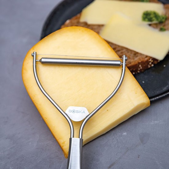 Een zilverkleurige Boska kaasschaaf met een gebogen handvat ligt op een puntige kaas. In de achtergrond een boterham met kaas.