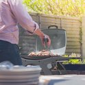 Man met tang grilt vlees op een grijze KitchenBrothers tafelbarbecue in de tuin.
