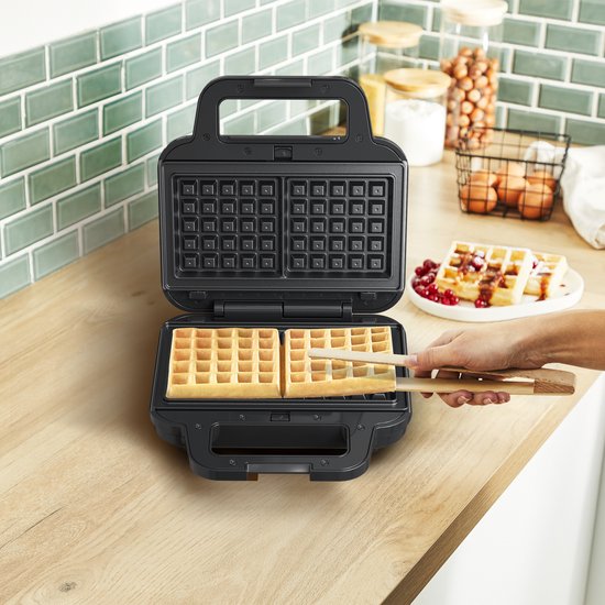 Zwart wafelijzer met twee vierkante wafels, in gebruik. Houten tangen verwijderen een wafel.