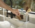Hand pakt een bruine spons uit een zilverkleurige gootsteen organizer naast een kraan met sop.