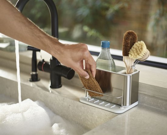 Hand pakt een bruine spons uit een zilverkleurige gootsteen organizer naast een kraan met sop.