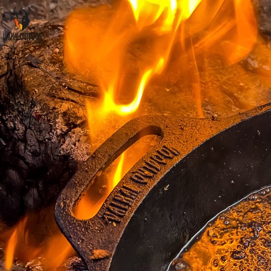 Gietijzeren koekenpan met een ingegraveerd Viking-logo bij het handvat, terwijl deze boven een kampvuur wordt verwarmd.