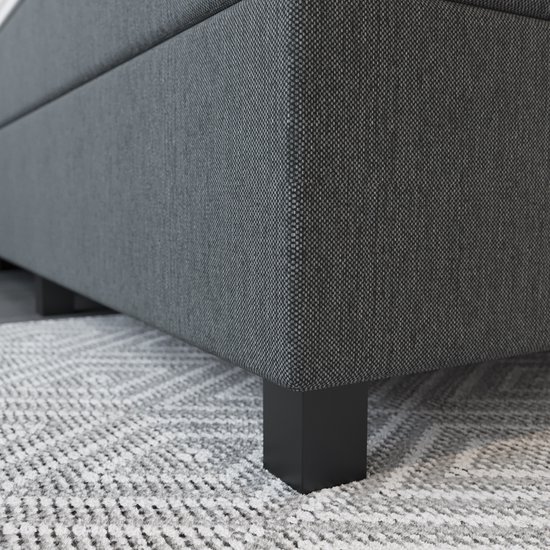 Detail van de grijze stof en zwarte poot van een boxspring, op een vloerkleed met ruitpatroon.