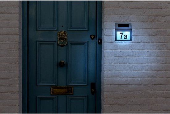 Zilverkleurige huisnummerlamp met nummer '7a' verlicht naast een blauwe deur, gemonteerd op een witte bakstenen muur.