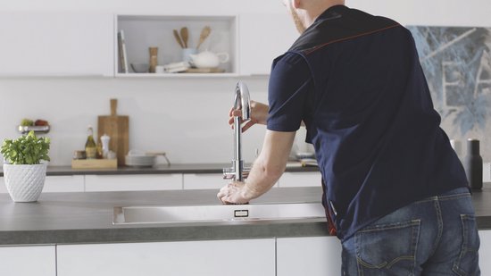 Monteur installeert chroom GROHE kraan in een moderne keuken met witte kasten en een metalen gootsteen.
