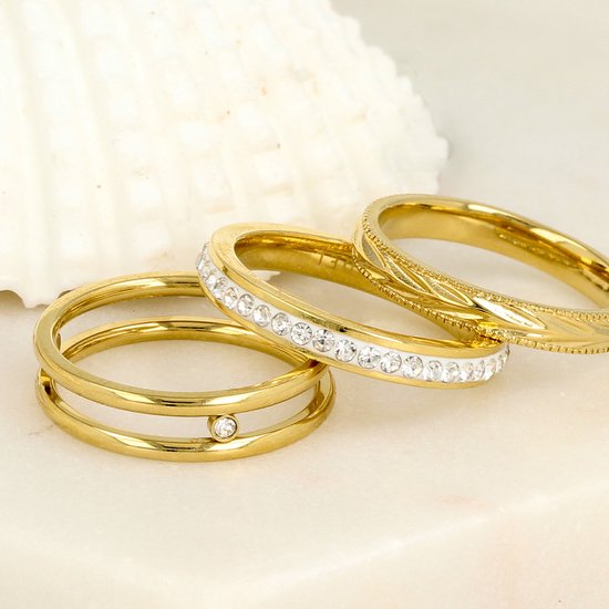 Drie gouden ringen op een marmeren achtergrond: een dubbele ring met zirkonia, een band met zirkonia en een gedetailleerde band.